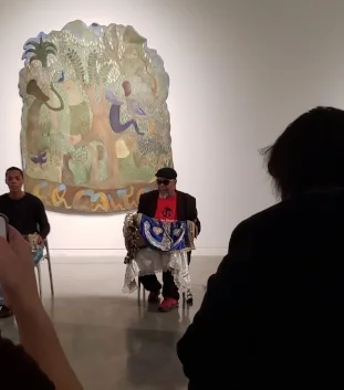 Two afro-cuban men play drums. The one on the left is younger and bit cut off by the frame. On the right is Roman Diaz, he is an older man, wearing a black berret, sunglasses, a red shirt and a black blazer. His large drum is horizontal across his lap and covered in a decorative blue, white and gold cloth. In the foreground are audience members, one holding up a phone to film. In the background is artwork by Manuel Mendive on a white museum wall. The art has a jungle like scene where humanoid figures are sitting on, around, and underneath a tree.