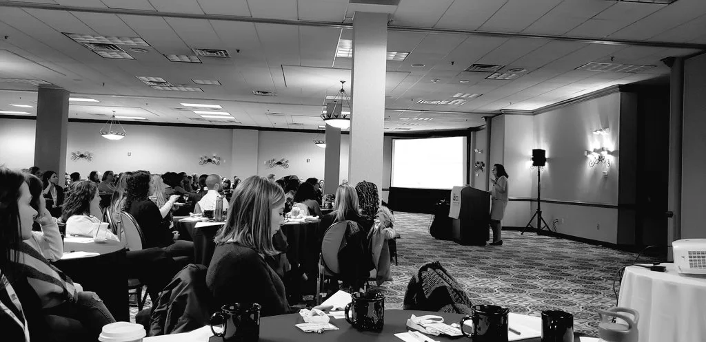 Black and white photo. Alicia F. Lieberman, a senior woman, addresses a large crowd of people from behind a podium. Behind and around Lieberman conference equipment can be seen including a large presentations screen, projector, speakers, and a banner with logo draped over the podium itself.