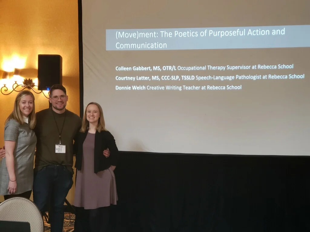 From left to right: Colleen Gabbert, young white woman. Donnie Welch young white man. Courtney Latter young white woman. All dressed professionally. The three are posing in front of their title slide on a large presentation screen with a black, curtain skirt. Behind them can be seen a speaker and light as well as the navy-yellow rug and yellow wall of the hotel ballroom.