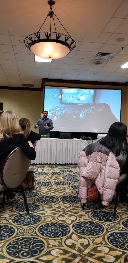 Chris Hernandez a young Puerto-Rican man, presents from behind a table to a group of people sitting. His body language suggests he is engaged and mid-speech. Behind him is a large presentation screen with “So…What is a Video Game” written over the image of a console controller. Around Hernandez can be seen equipment such as a laptop, sound board, and projector. The presentation is taking place in the hotel ballroom and the navy and yellow rug and yellow wall of the room are seen in the photo.