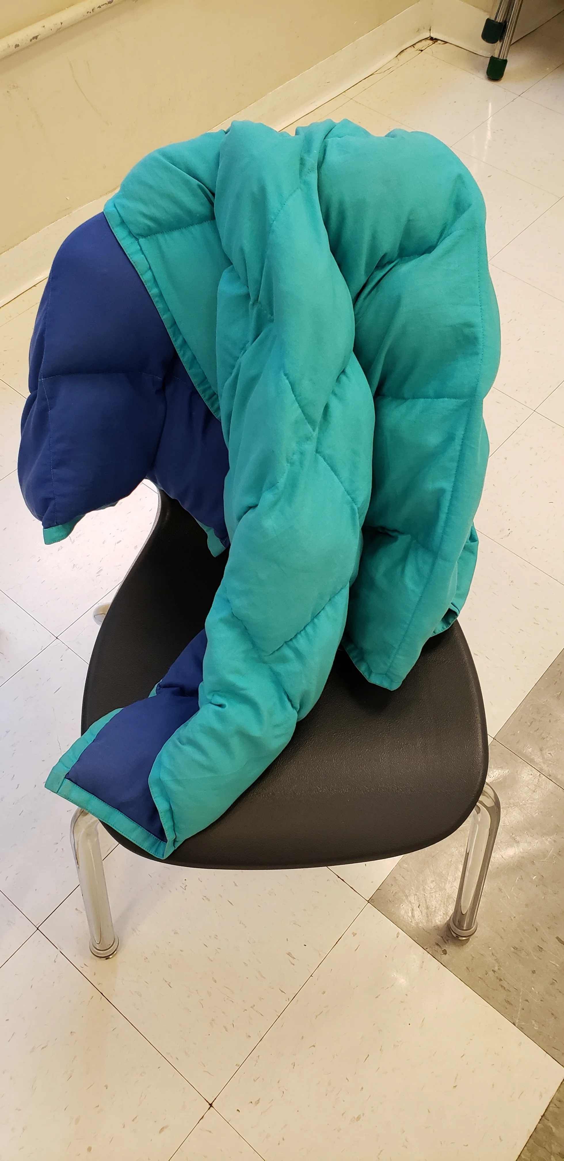 Teal and dark blue weighted blanket sitting on top of a black chair in a tiled classroom.