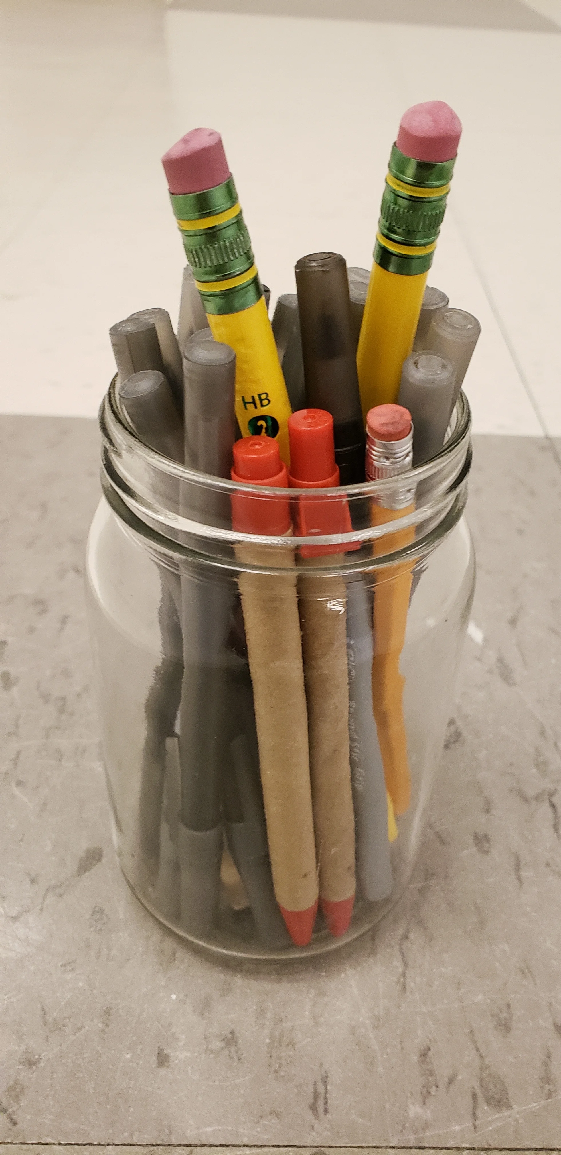 Clear mason jar of writing utensils on a tiled classroom floor. The Jar has plastic ball point pens with both smooth and rough grips, pens with cardboard wrapping, regular sized pencils, and giant pencils.