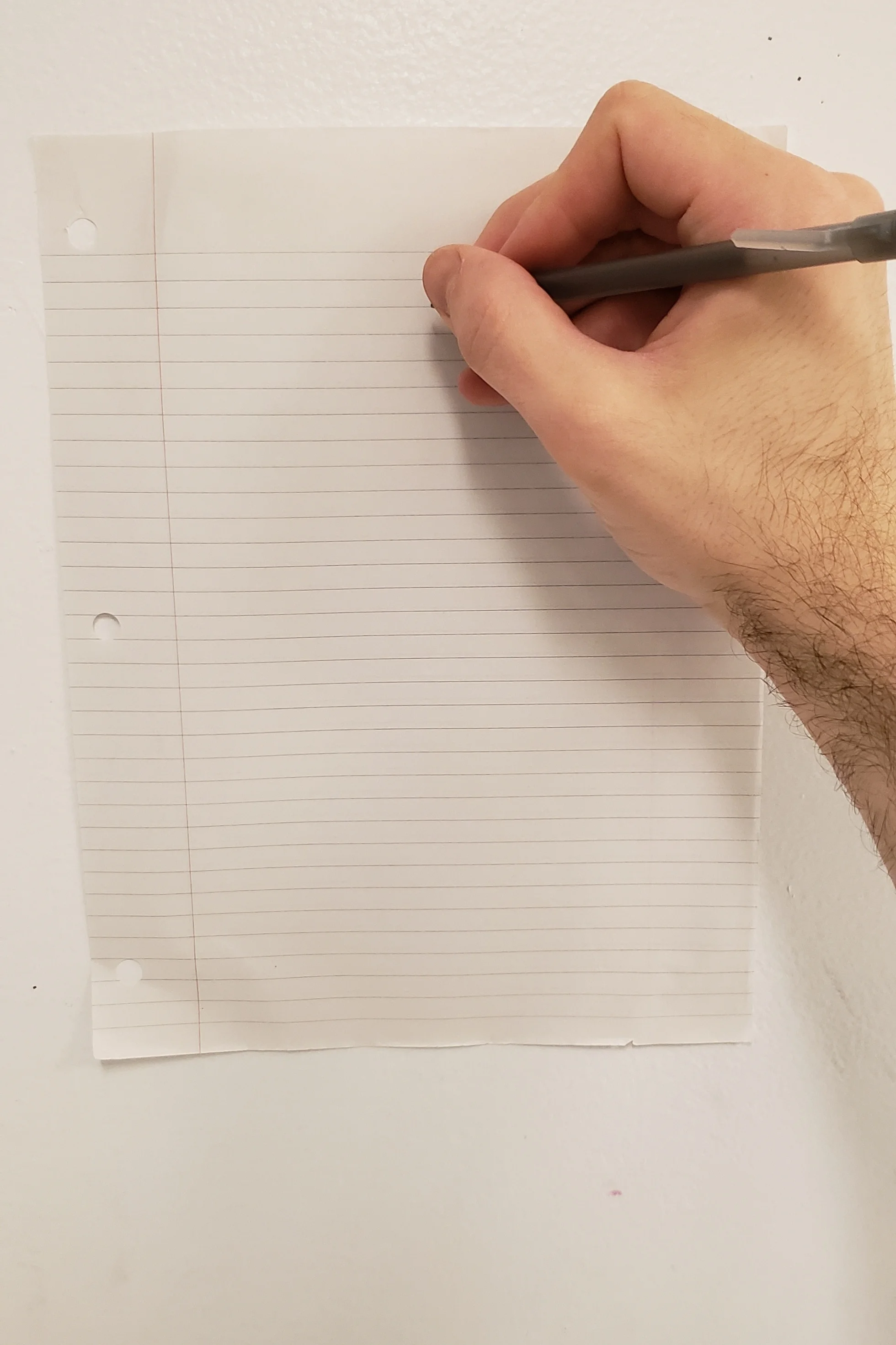 A hand holds a pen to a piece of lined paper taped to a white school wall.