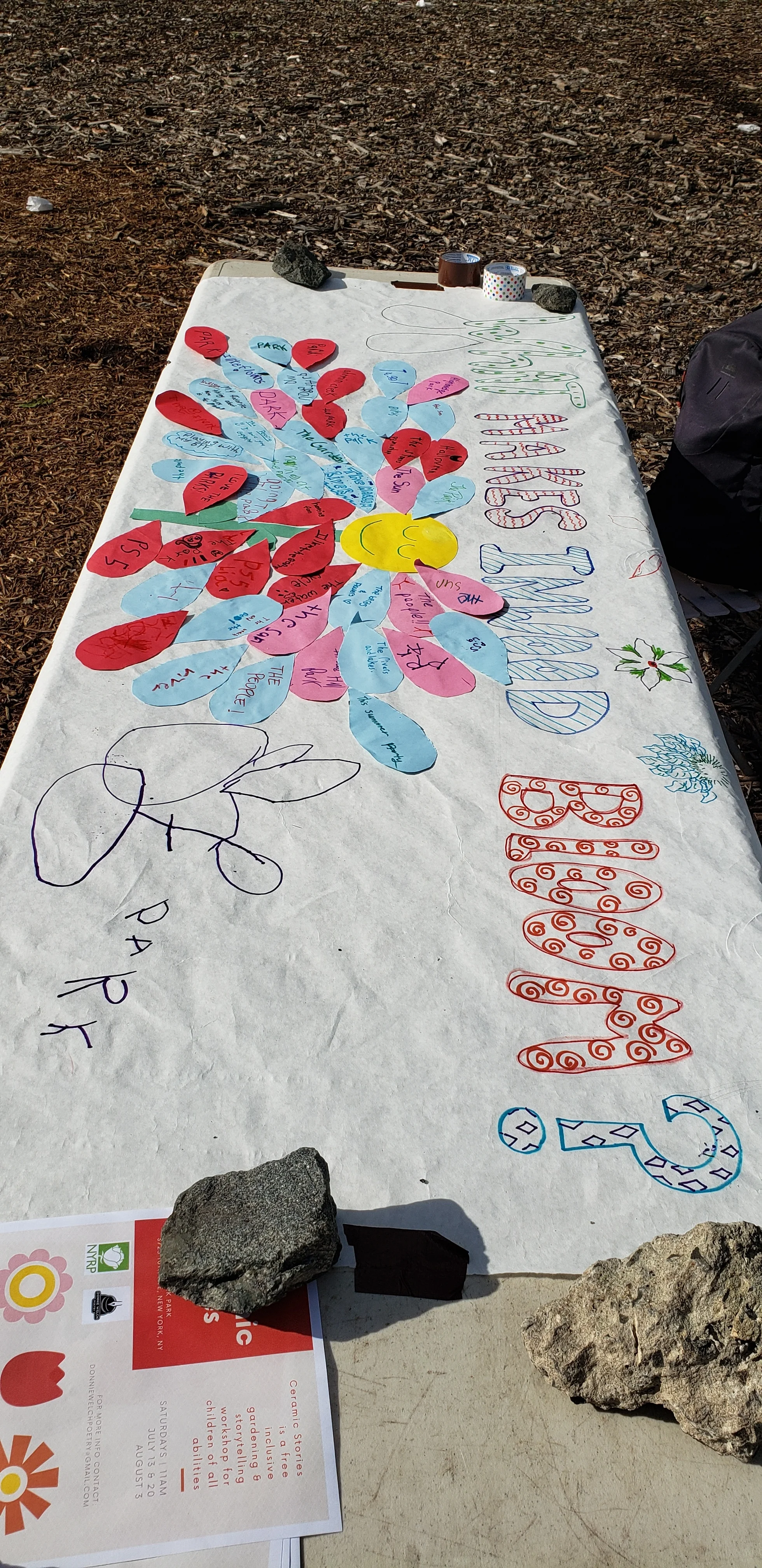 Photo of art mural on fold out table. The mural is on a long white sheet of paper and covered with “flower petals” in red, teal, and pink all with writing on it that is indiscernible at this distance. “What makes Inwood Bloom” is written in multi-colored large letter up top. On the mural can also be seen some writing and doodles. The table has some rocks on it, holding the paper, supplies, and other objects down in the wind.