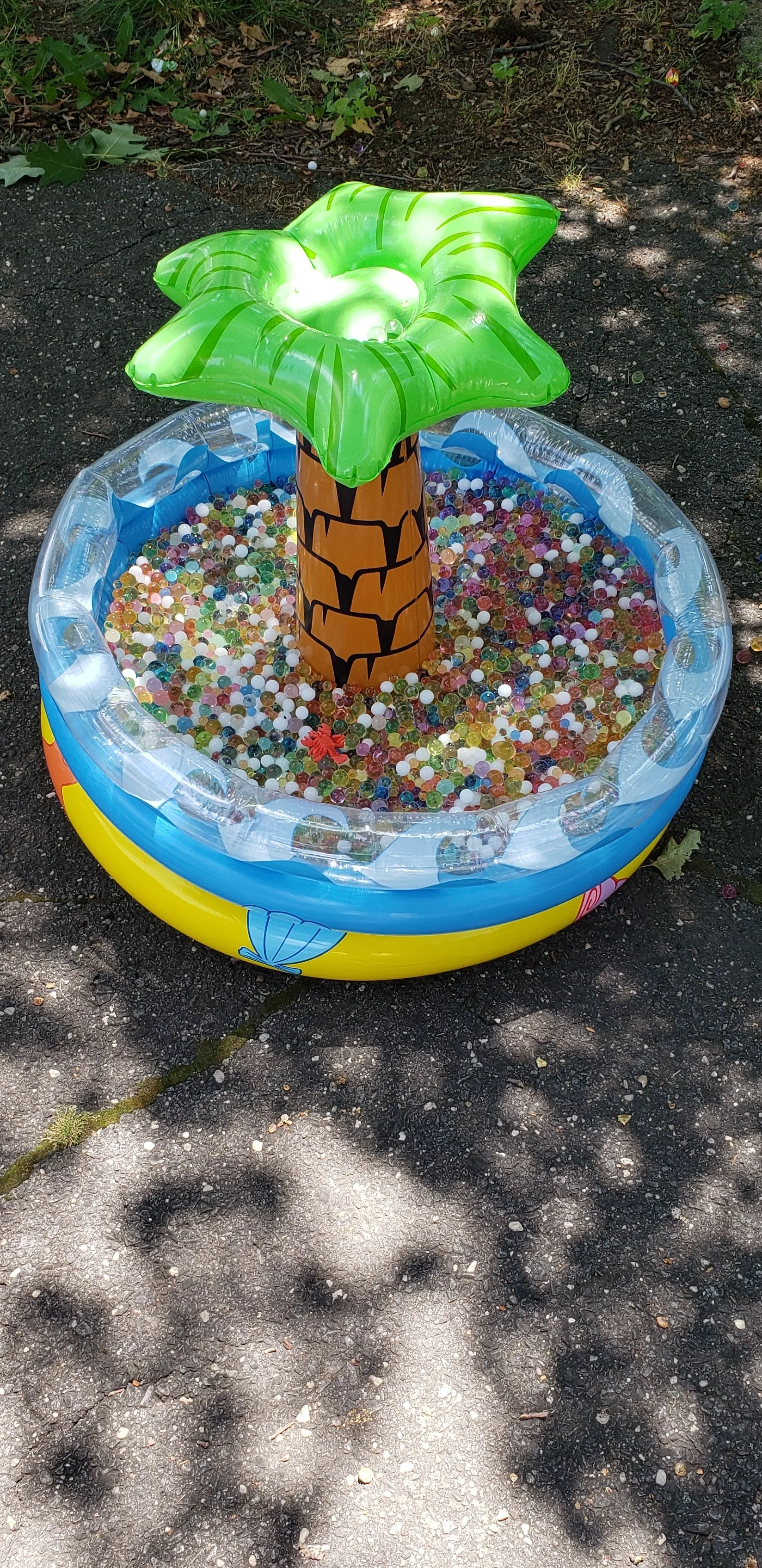 Inflatable, plastic swimming pool with an inflatable palm tree in the middle of it situated on a piece of asphalt. The pool is filled with water-bead sensory materials.