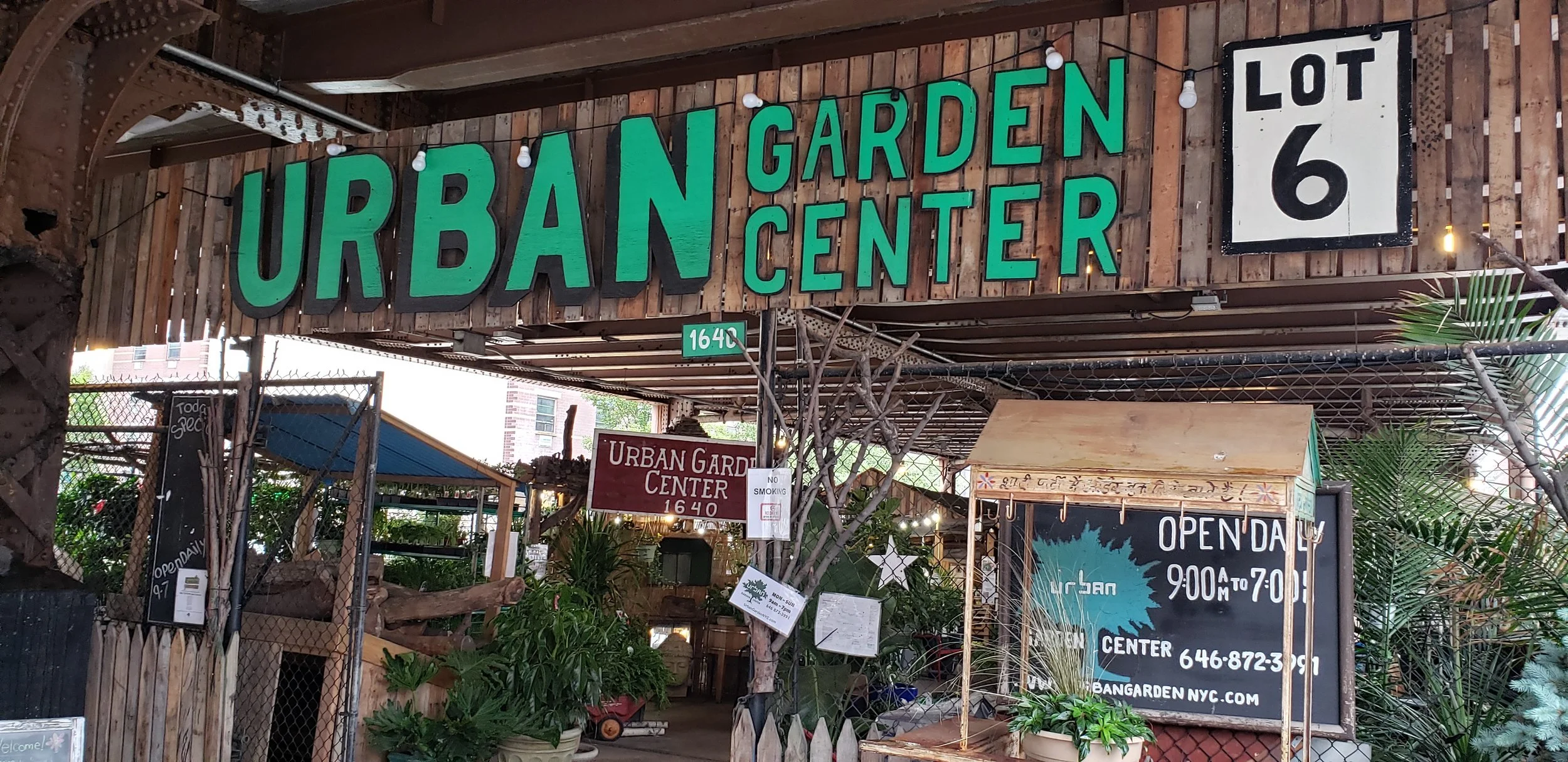 Entrance to the store “Urban Garden Center” The store's name is in large green block letters on a wooden frame with the word “Urban” being the largest. In the background can be seen plants and garden supplies such as fencing as well as signs denoting the stores hours and sales.