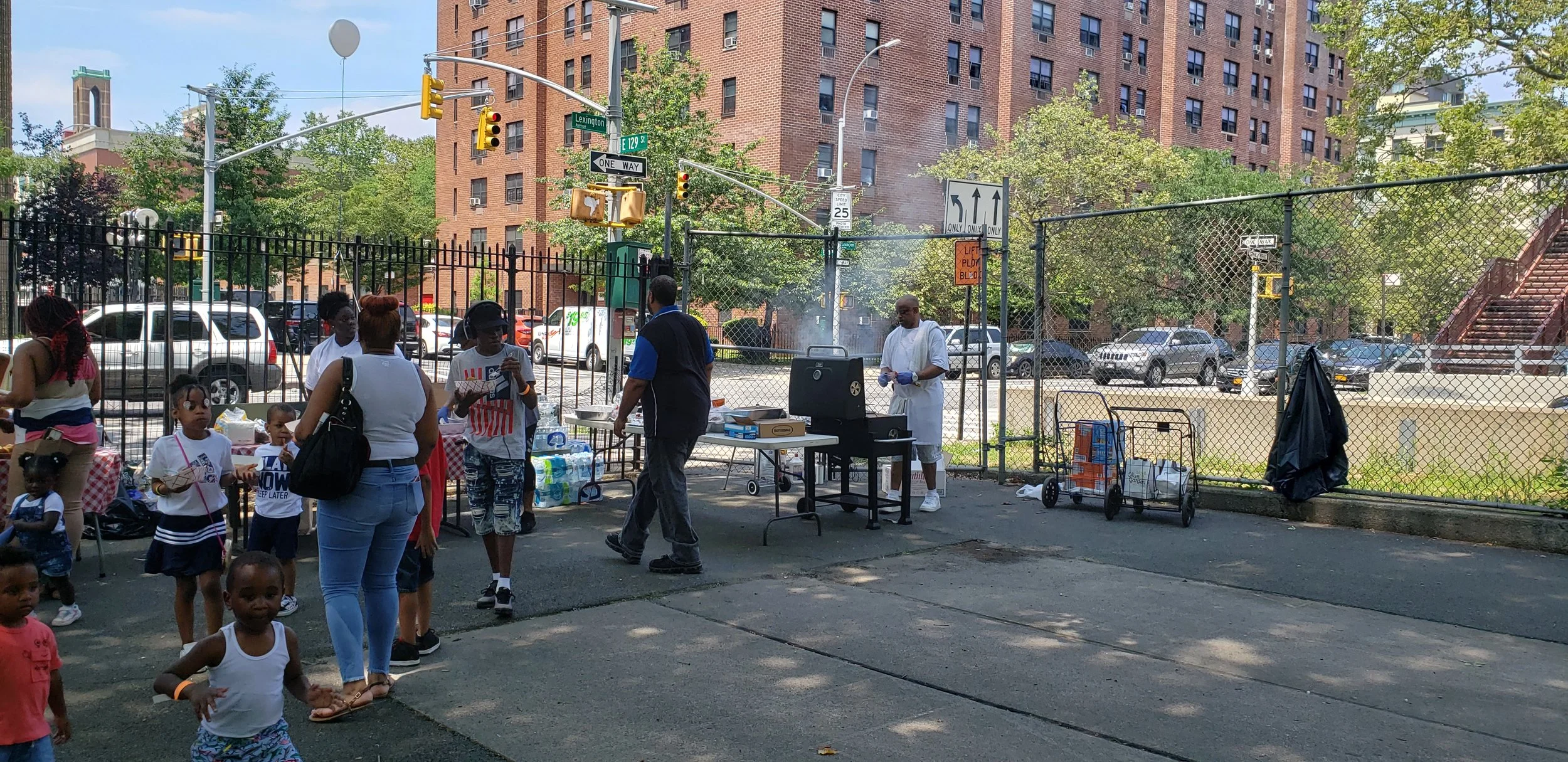 Autism Family Fun day grill set up, families and children are in the foreground of a shot of a food line. Tables in the background have the food on them and in the top right of the photo the grill master cooks while smoke can be seen coming off the grill.