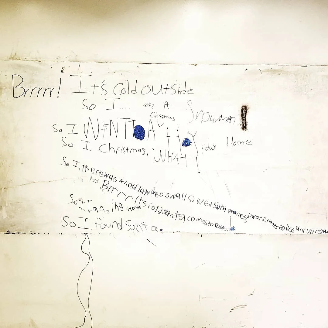 Words on a white board in black expo marker read “Brrrr! It’s cold outside so I…” with various student answers written. The answers are hard to completely discern at the distance of the photo.