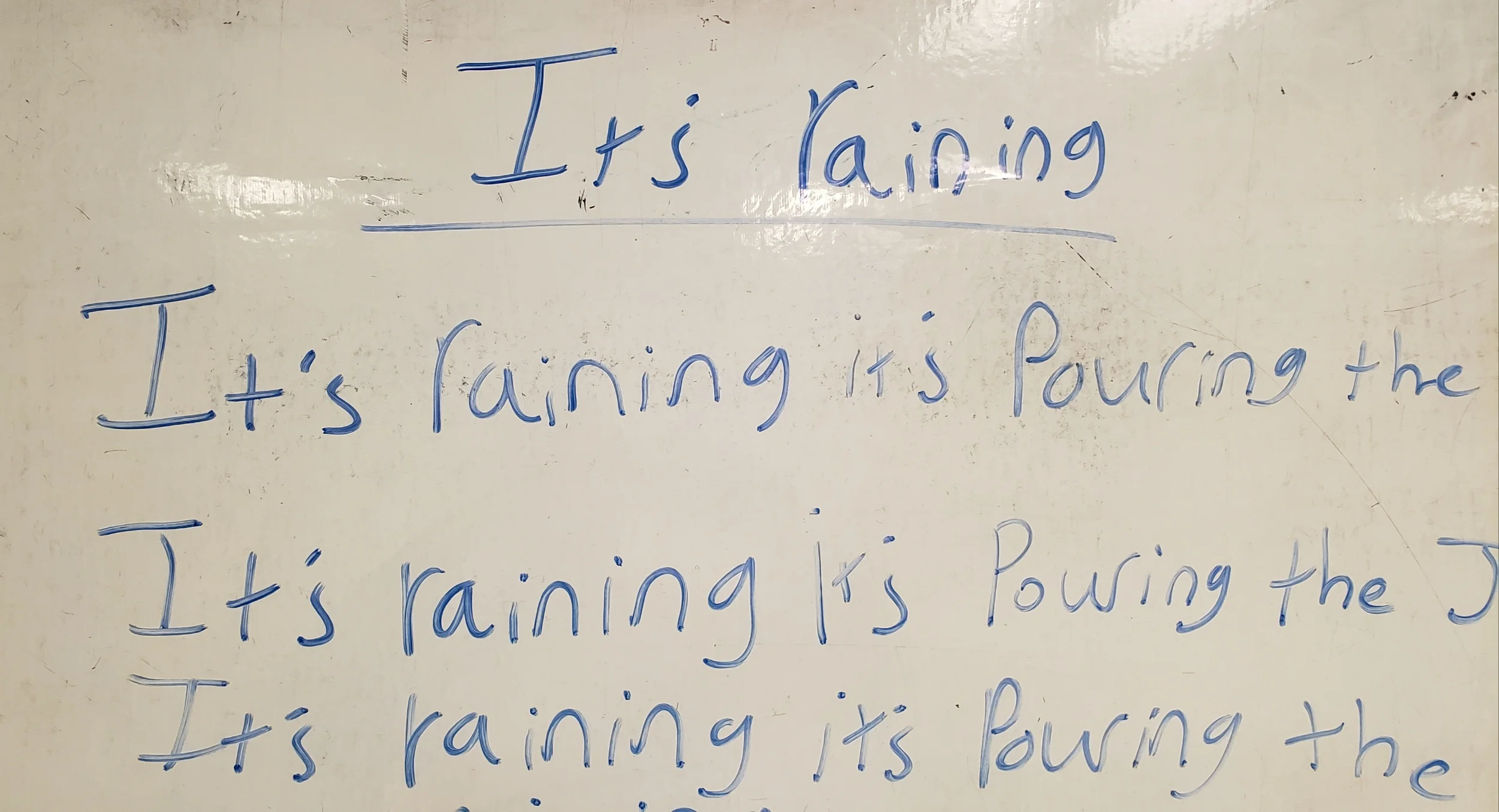 Words on a white board in blue expo marker read “It’s raining it’s pouring the” This line is repeated again and again.