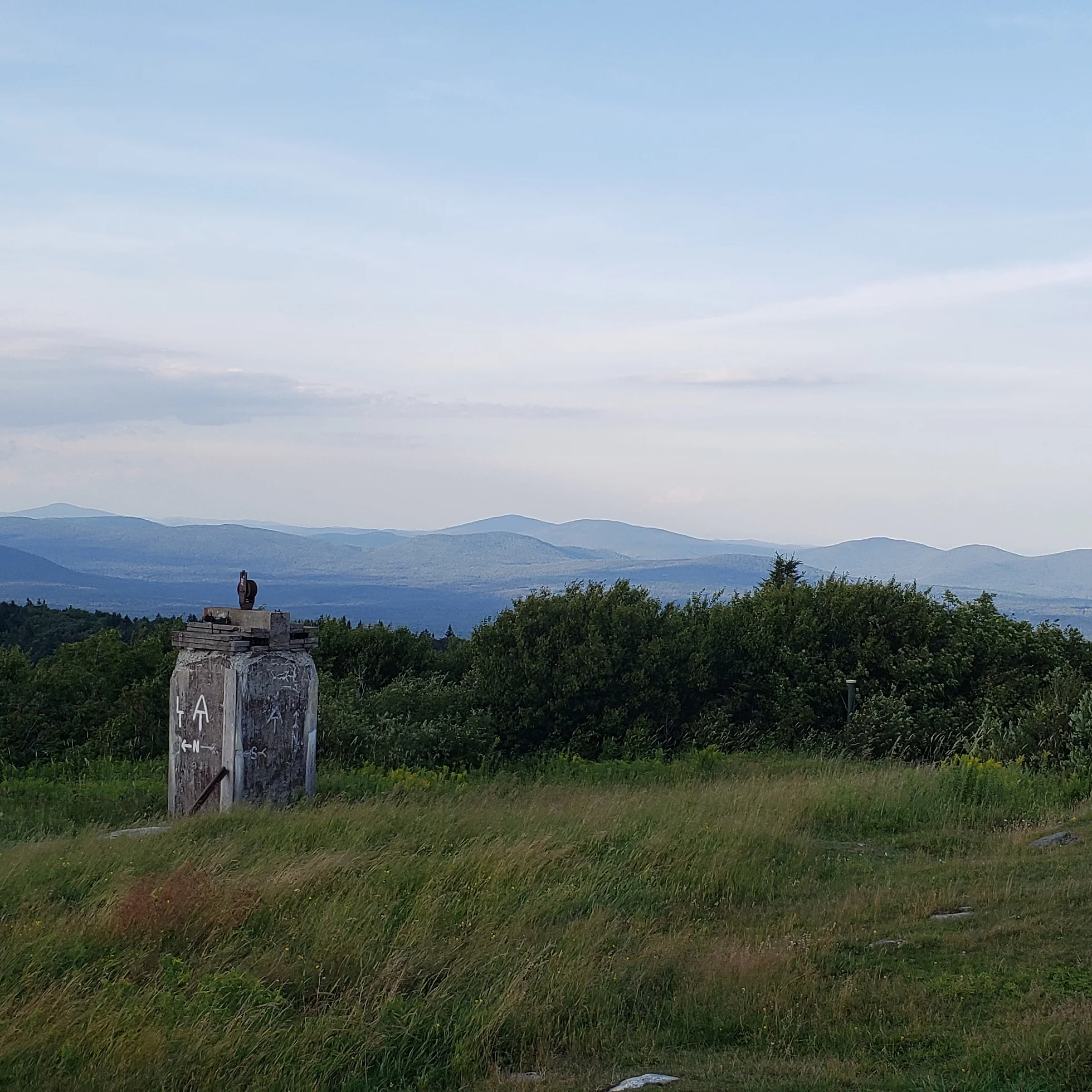 Sunset view off Bromley Mountain. In the foreground is a field of tall grass bent by the wind and an old stone work with LT and AT symbols and white N for North painted on in white.In the background can be seen ranges of mountains pale blue and purple from the light at sunset.