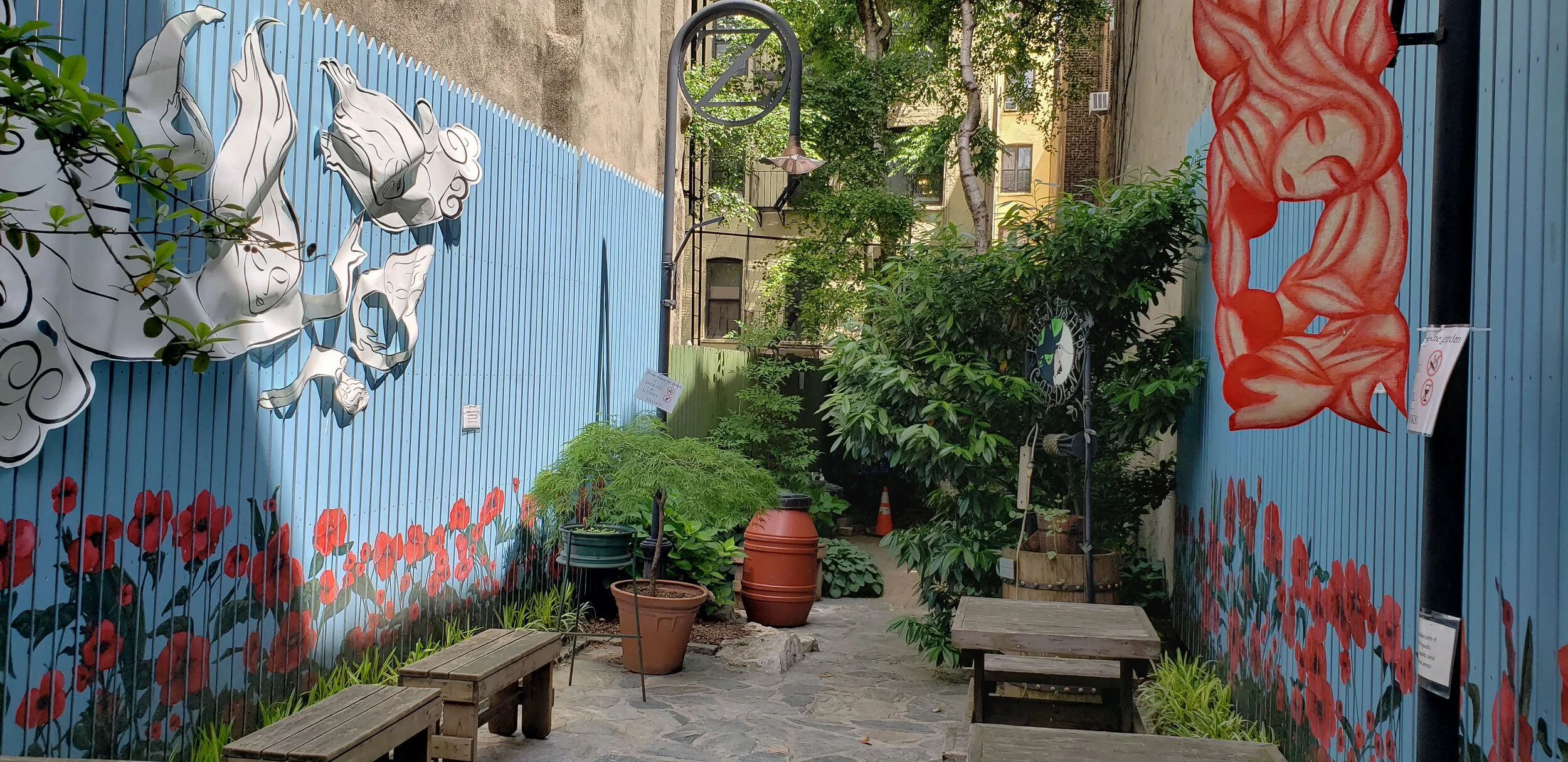 Community garden tucked between tall New York City buildings. In the foreground can be seen pavers marking a path, benches, compost bin, rain bucket, and array of trees and bushes. The side fencing of the garden is painted teal with red, poppy flowers painted eye level. Above are hung red and white cloud-human figures from a recent public-art exhibit.