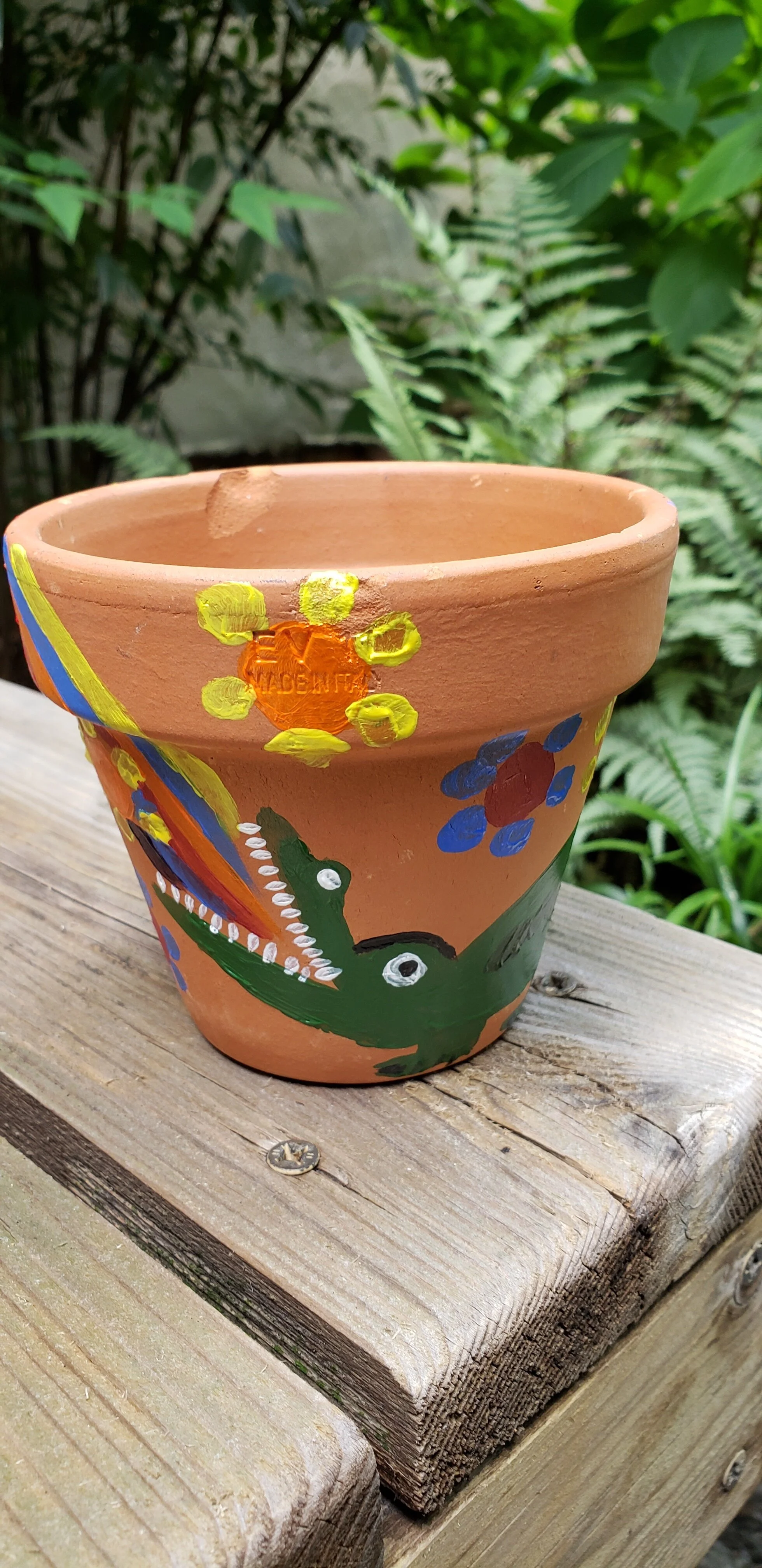 Terra Cotta flower pot with a painted green alligator. The alligator has a rainbow coming out of its mouth and flowers around it. The pot is sitting on a wooden bench and in the background is the greenery from a bush and ferns.