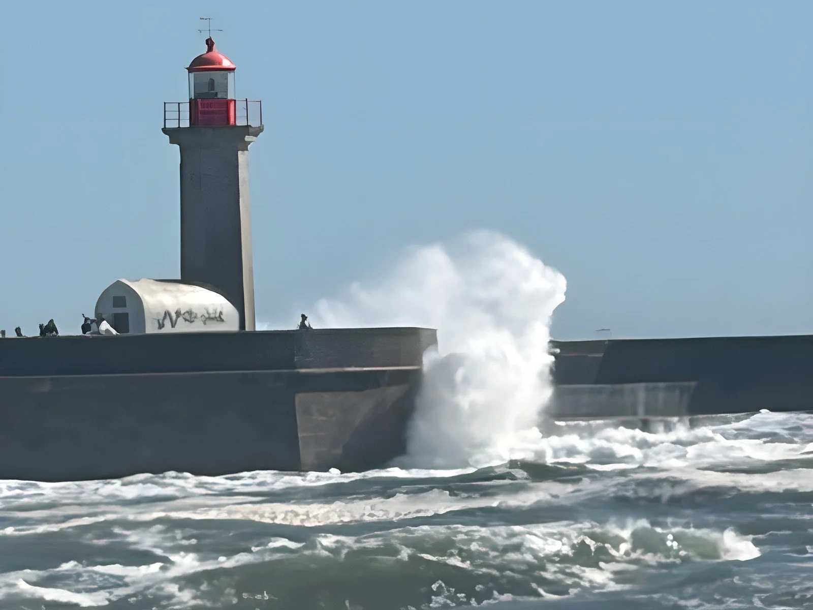 A wave crashing against a harbour wall beneath a lighthouse, standing steady in rough seas