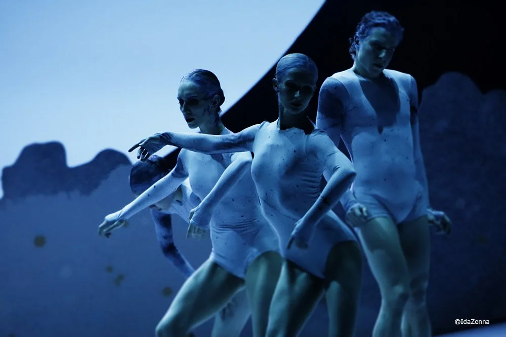 Four dancers in white costumes perform on stage against a dark background with blue lighting.