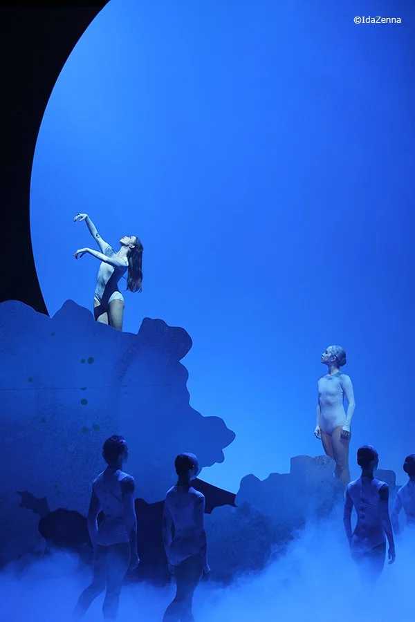 The image depicts a stage performance with dancers under blue lighting. Two female dancers are elevated, one with her head tilted backward and arms raised, and the other standing on the stage looking up. Several other dancers are standing below, partially obscured by fog, creating a surreal, aquatic-themed scene.