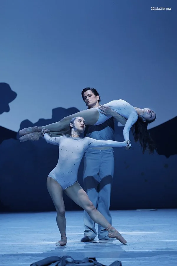 Ballet dancers performing on stage with a male dancer lifting a female dancer horizontally as a third dancer supports her legs; blue lighting and backdrop