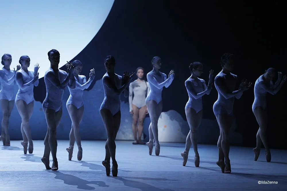 A ballet performance with a group of dancers on stage, lit with blue lighting, with one dancer in the background in white attire.
