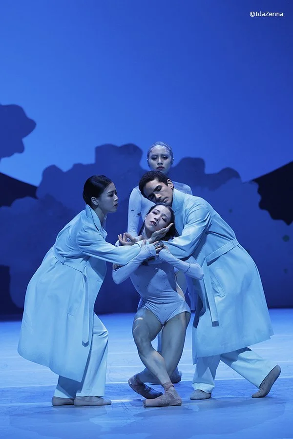 Four ballet dancers perform on stage in blue costumes, with three adults and one child, against a blue background.