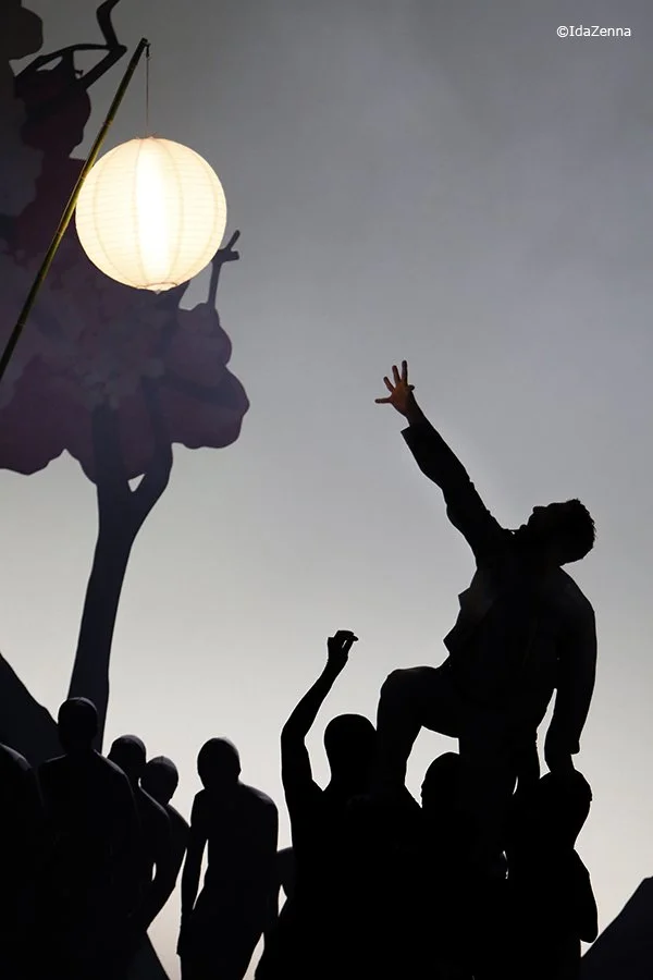 Silhouette of a person being lifted on others' shoulders, reaching upward, with a paper lantern hanging from a pole against the sky, creating a scene reminiscent of a festival or celebration.