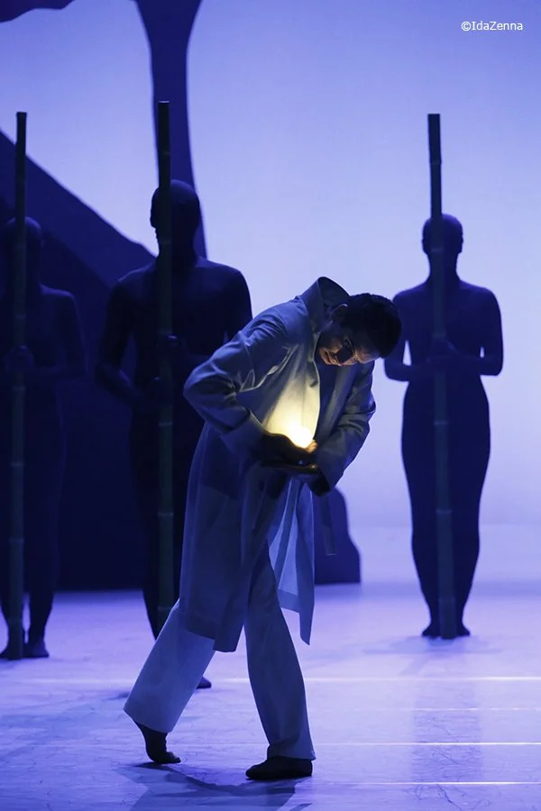 A person in a white coat stands on stage, looking at a glowing device in their hands, surrounded by four silhouetted figures holding spears against a blue background.