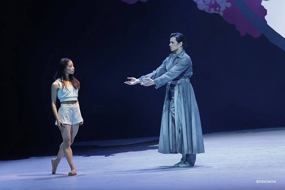 A male performer and a female performer are on stage in a dance performance, with the male extending his arms towards the female who is standing with her hands at her sides, wearing dance costumes.