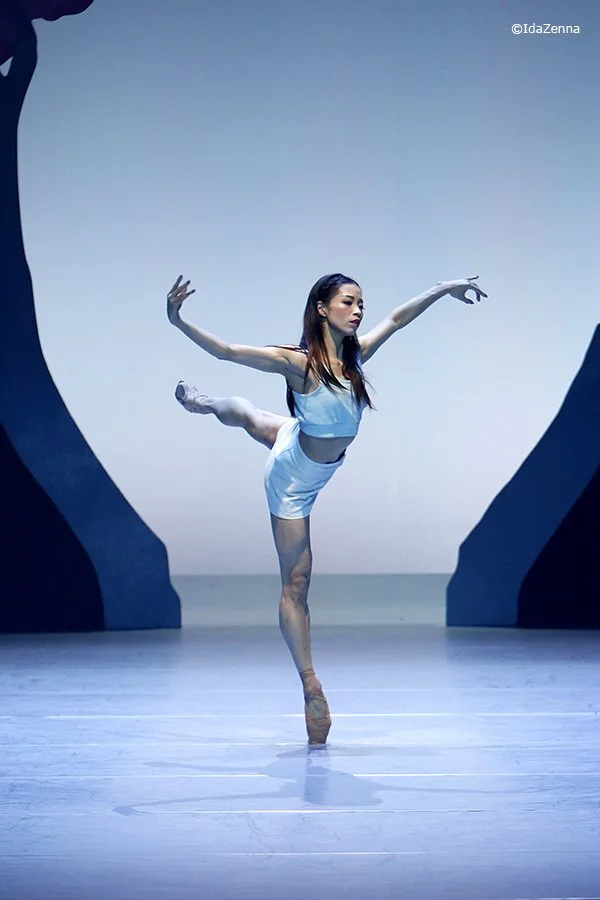 A female ballet dancer performing a pose in a dance studio with scenic set pieces in the background.