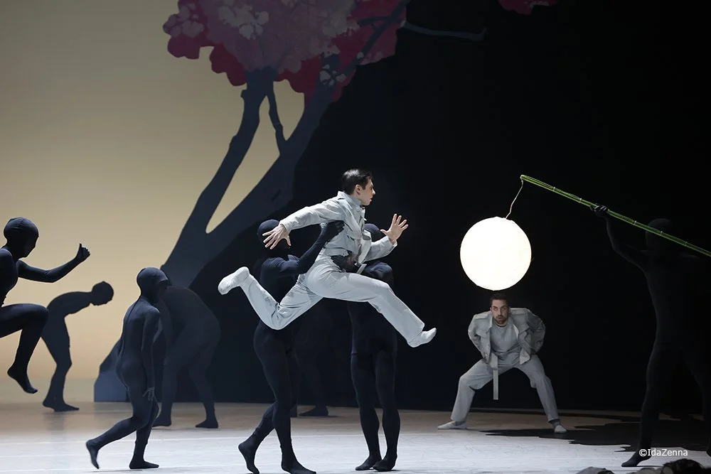 Performers on stage, some standing and others reaching or leaping, with a large white orb hanging from a pole and a backdrop of a tree with pink blossoms.