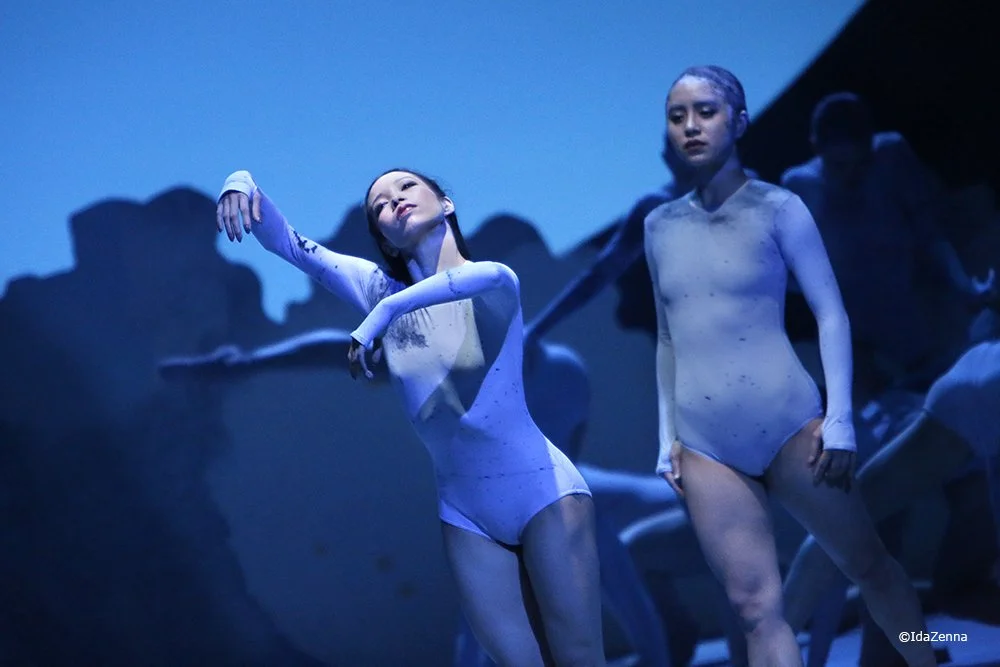 Two ballet dancers in white costumes performing on stage with blue lighting and shadows.