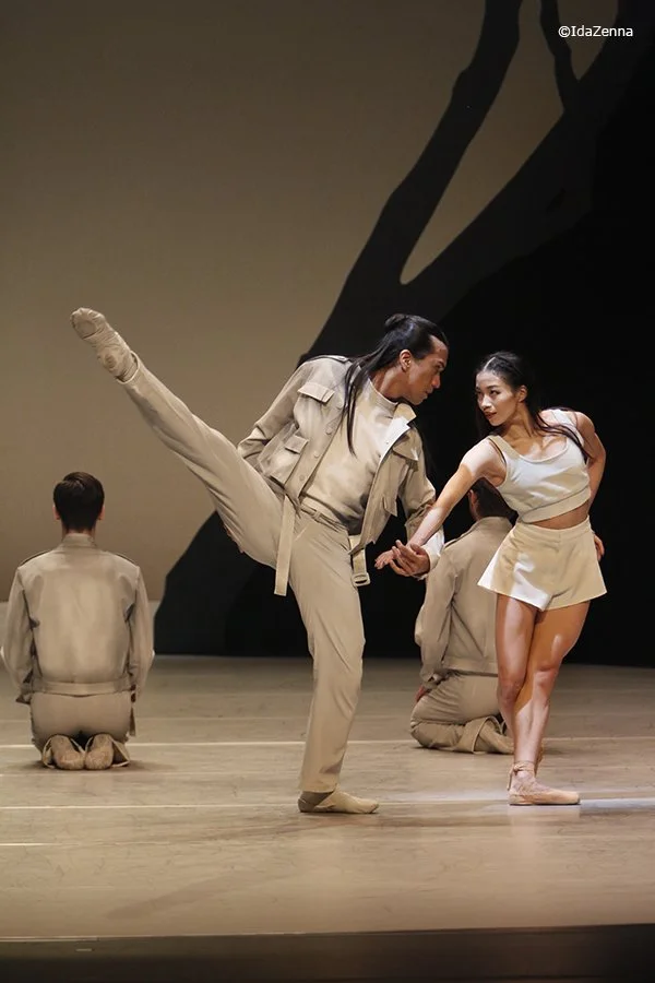 Two dancers performing on stage, one with a high kick and the other holding her hand, with two other dancers kneeling behind them.