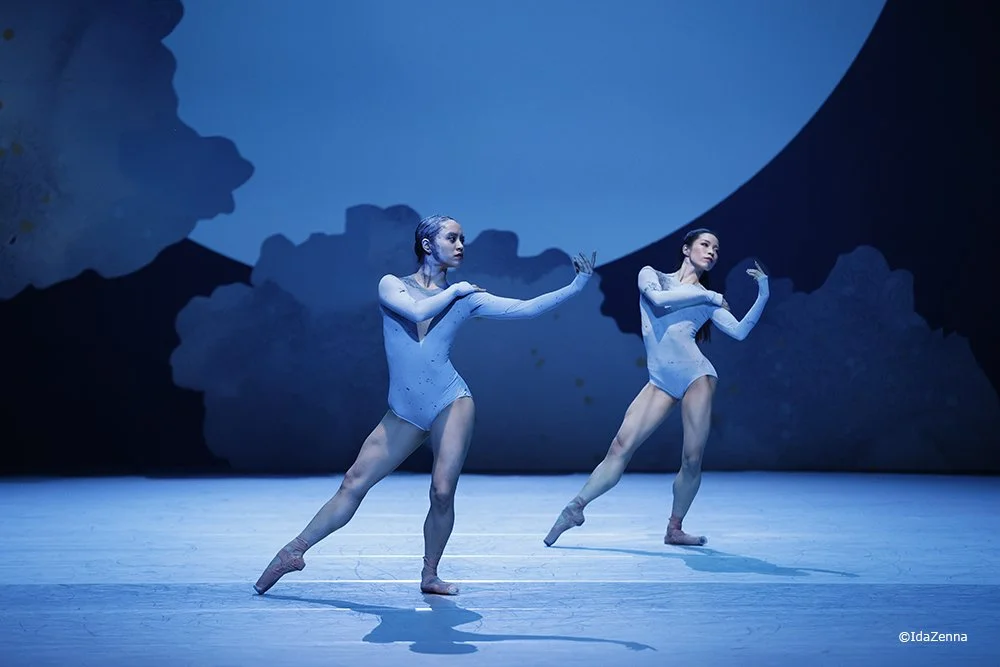 Two ballet dancers perform on stage with a blue and dark background, wearing light-colored costumes and ballet shoes.