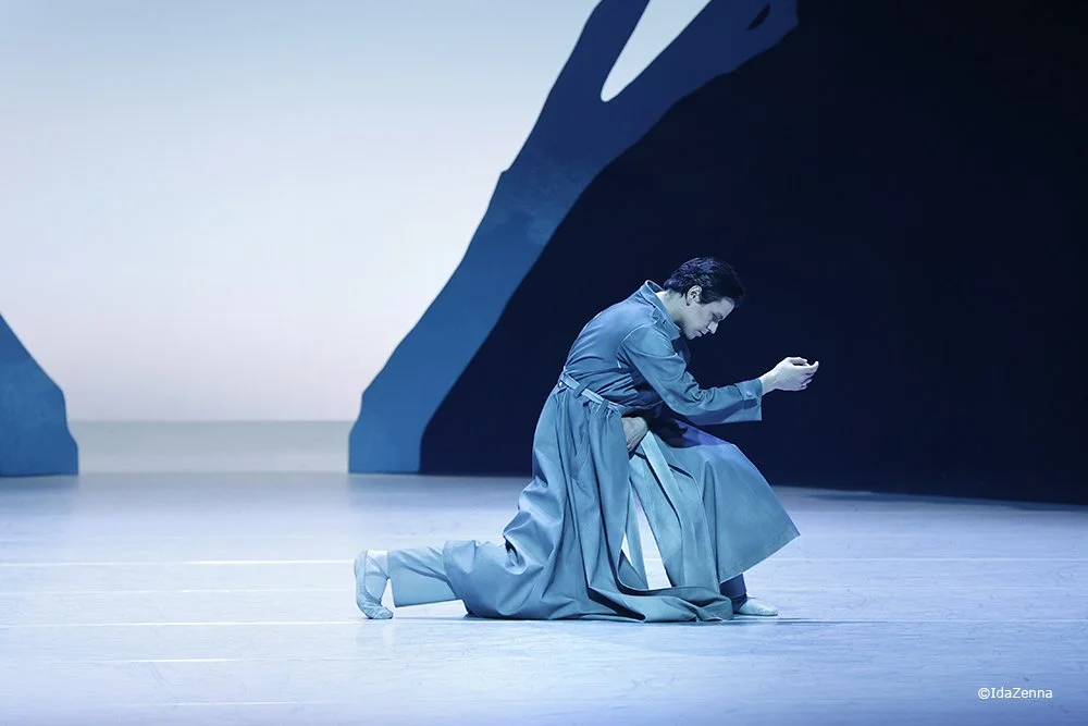 A person in blue clothing kneels on stage floor, looking at a device in their hand, with large dark blue structures behind them.