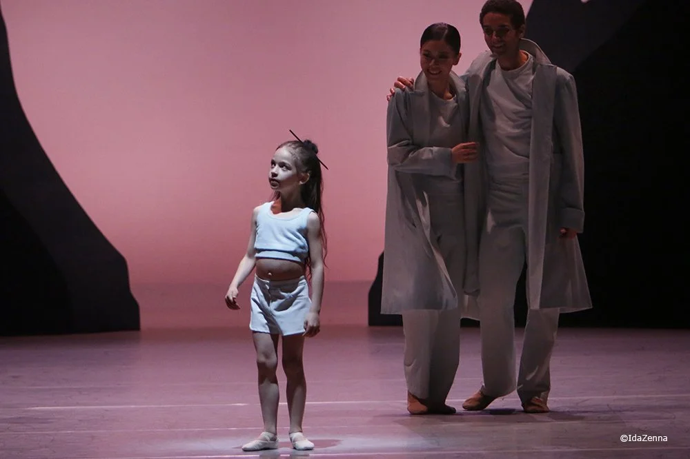 Three dancers on stage, with one young girl in a light blue and white outfit standing apart from an adult couple in gray costumes.