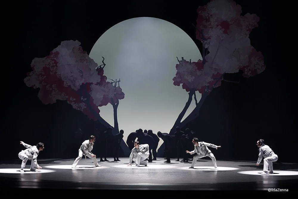 The image shows a stage performance with six dancers in white outfits performing in front of a backdrop depicting a large moon and pink cherry blossom trees.