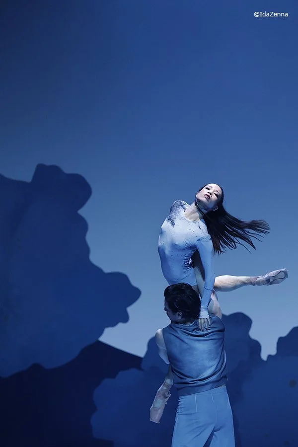A ballet dancer is being lifted by a male dancer on stage with blue lighting and shadowy mountain-like background.