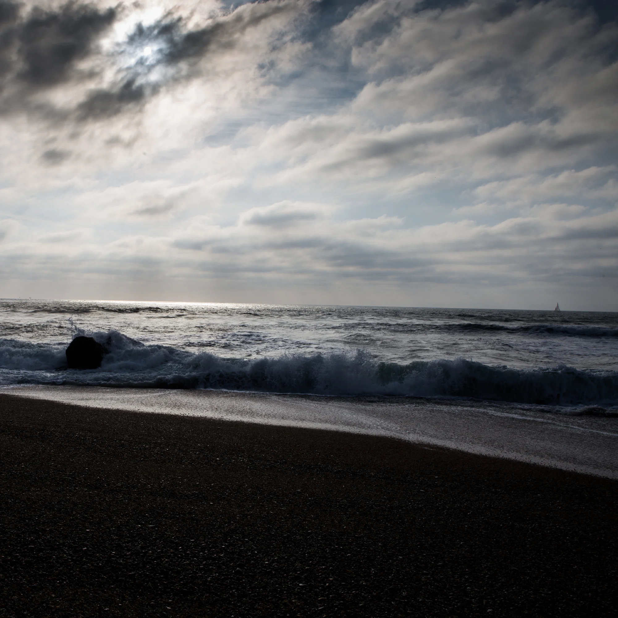 A cloudy beach scene with waves crashing on dark sand and a sailboat in the distance.
