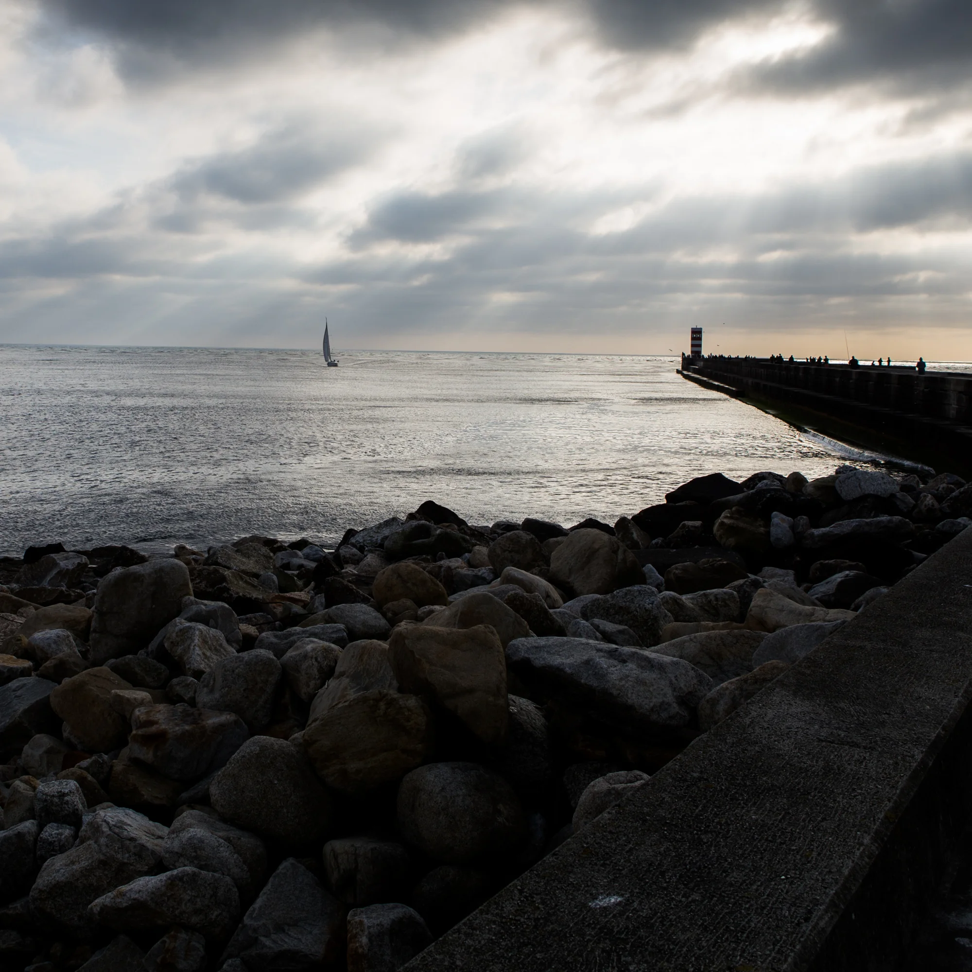 A rocky shoreline with a pier extending into the water, a lighthouse at the end of the pier, and a sailboat on the water under a cloudy sky.
