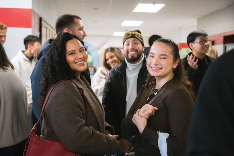 A smiling woman and man standing together indoors, with the man’s arm around the woman's shoulder. People are mingling in the background near a staircase and large windows.