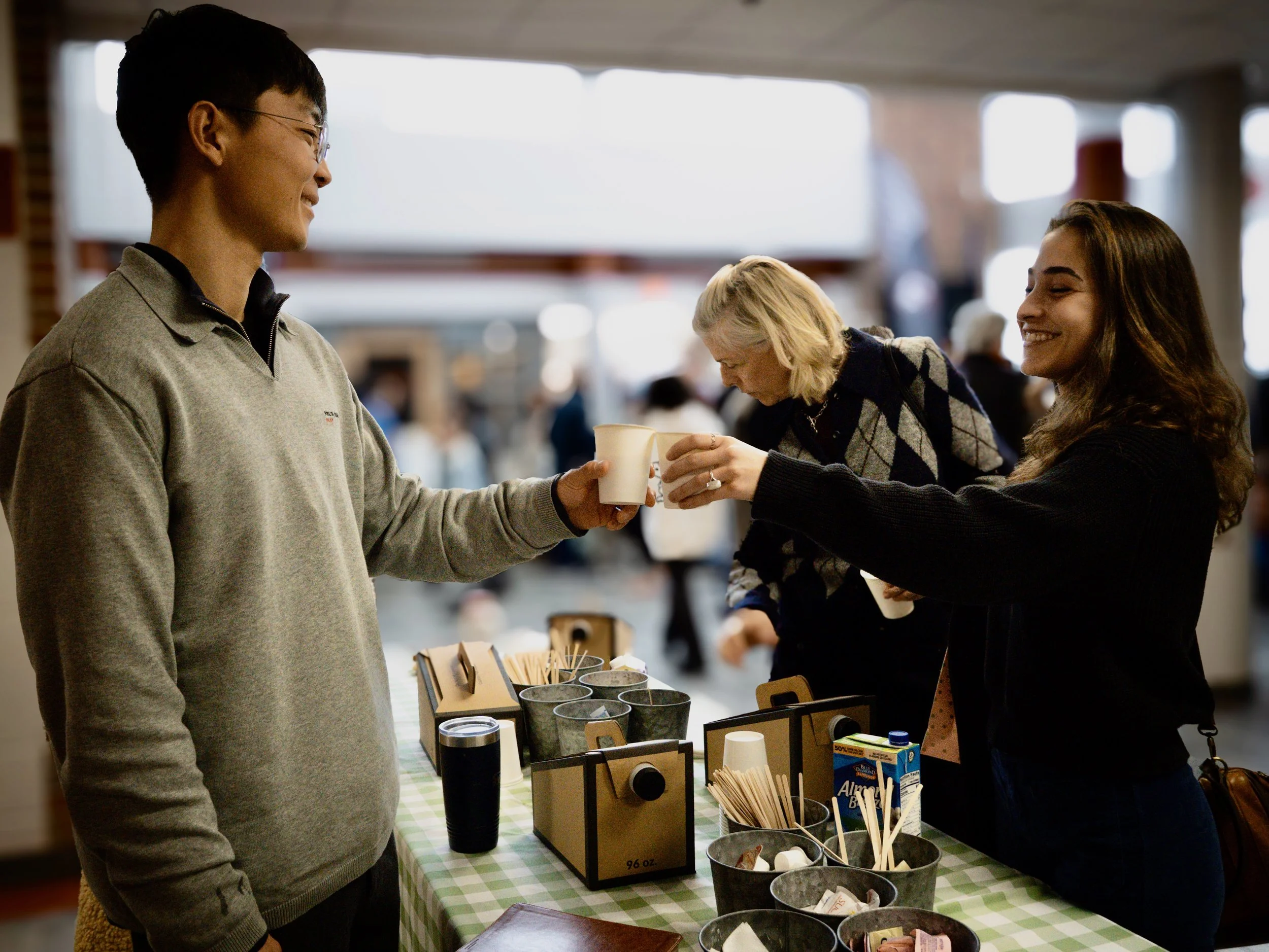 A smiling woman and man standing together indoors, with the man’s arm around the woman's shoulder. People are mingling in the background near a staircase and large windows.