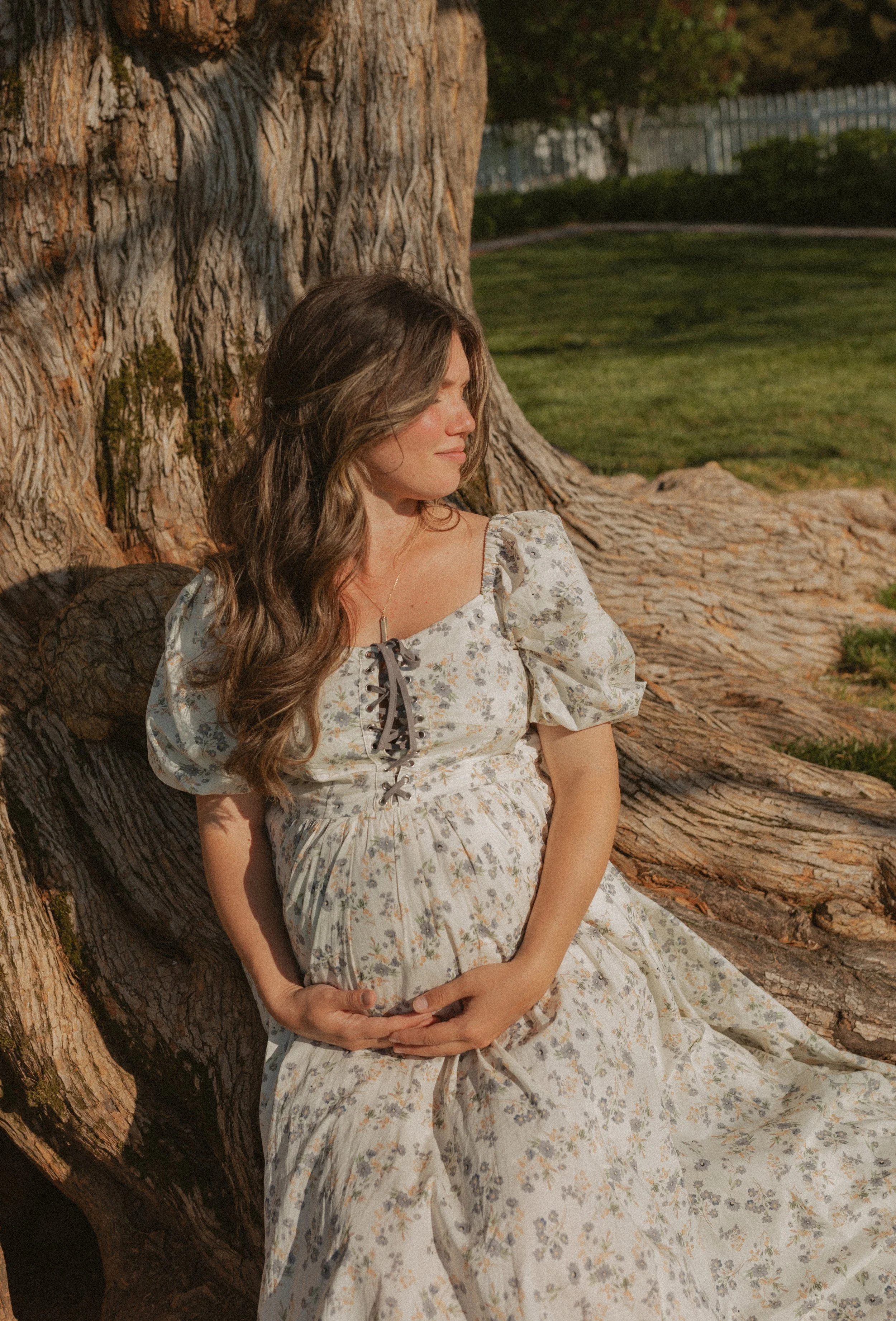 Pregnant woman sitting and leaning against a try while holding her hands under her bump and closing her eyes smiling