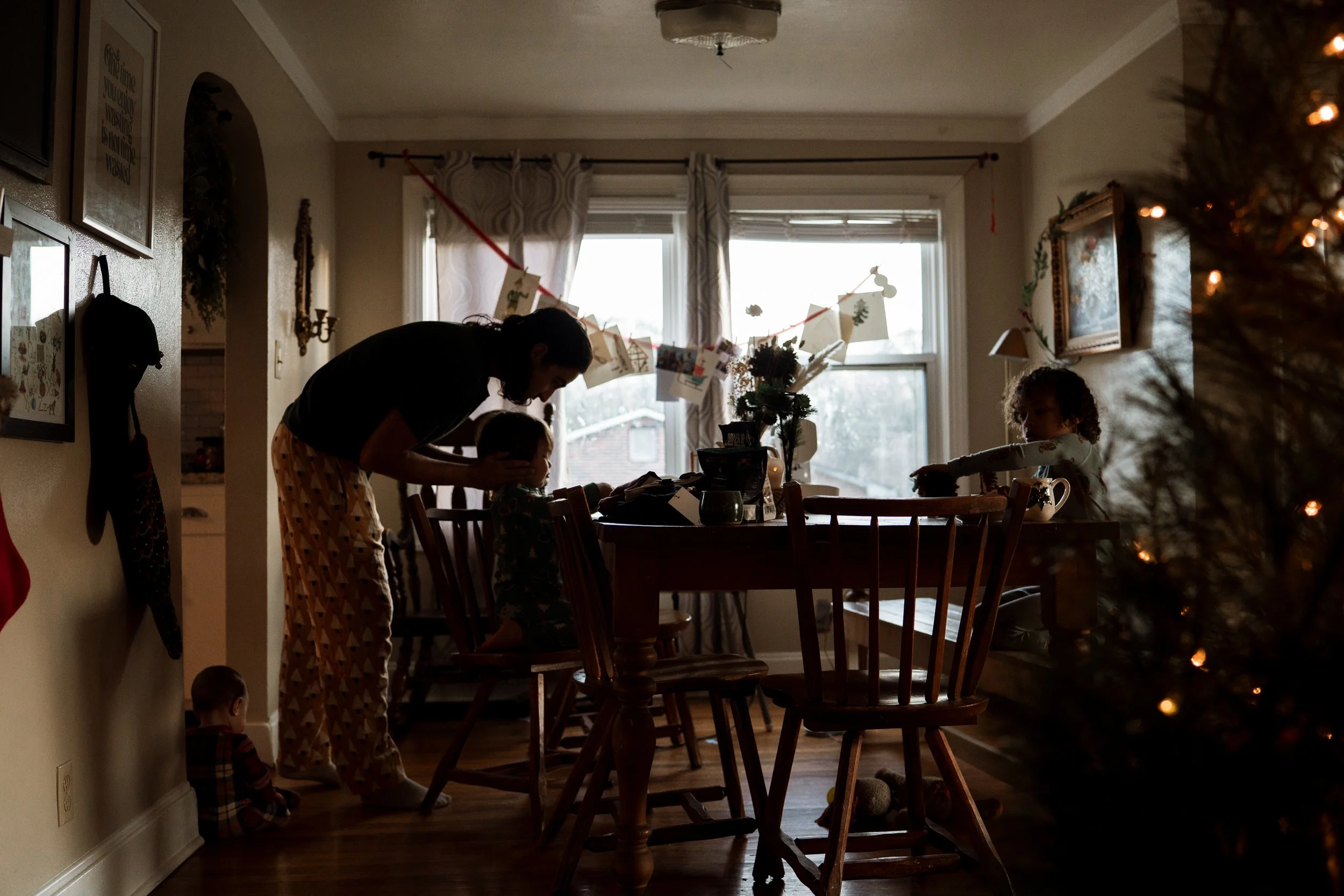 children sitting at dining room table in a Christmas themed home while dad reaches down to kiss one of the Childs head