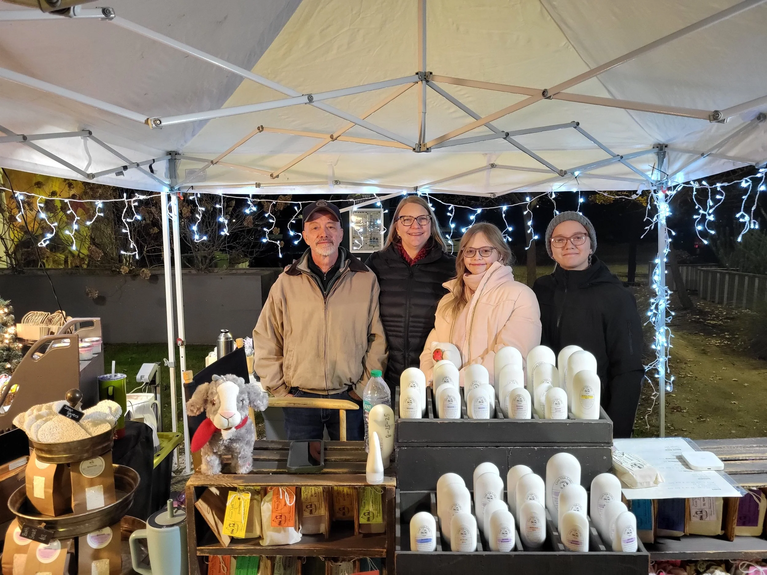 Group of four people standing behind a table selling various products at an outdoor market under a canopy decorated with string lights, with trees and darkness in the background.