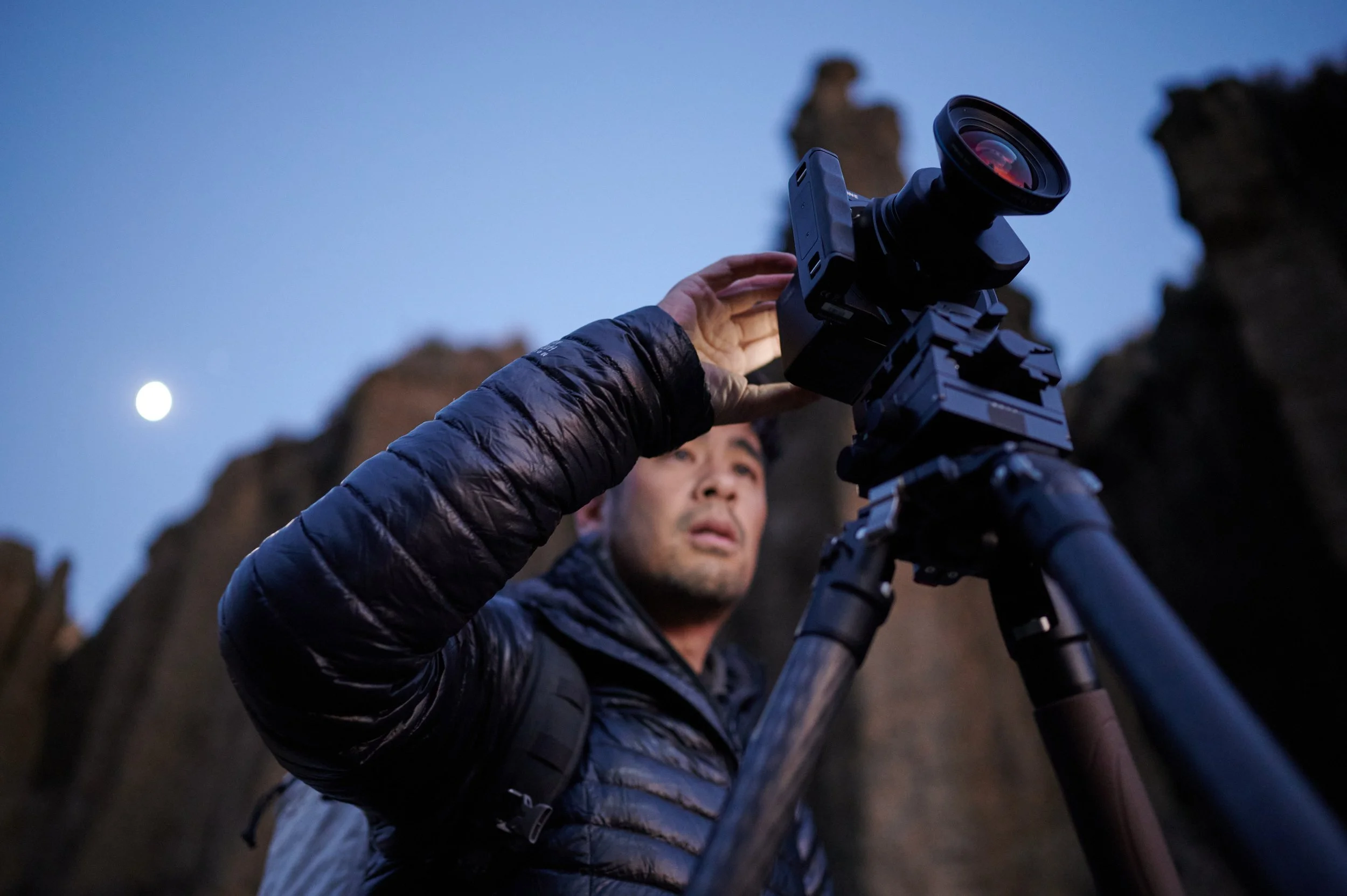 Man operating a camera on a tripod outdoors at dusk, with rocky cliffs and a visible moon in the sky.