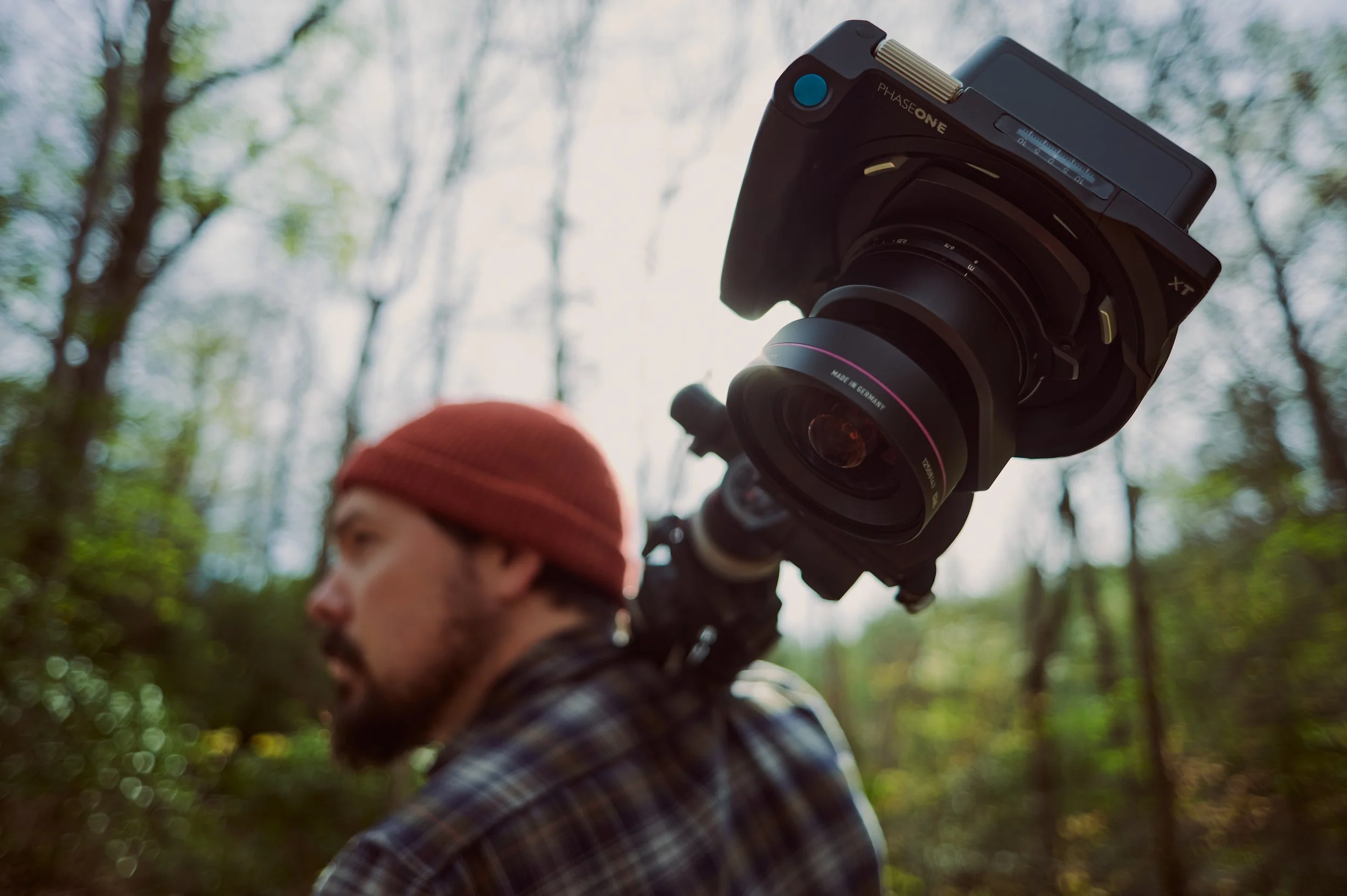 A man in a plaid shirt and red beanie is carrying a professional video camera on his shoulder in a wooded area.
