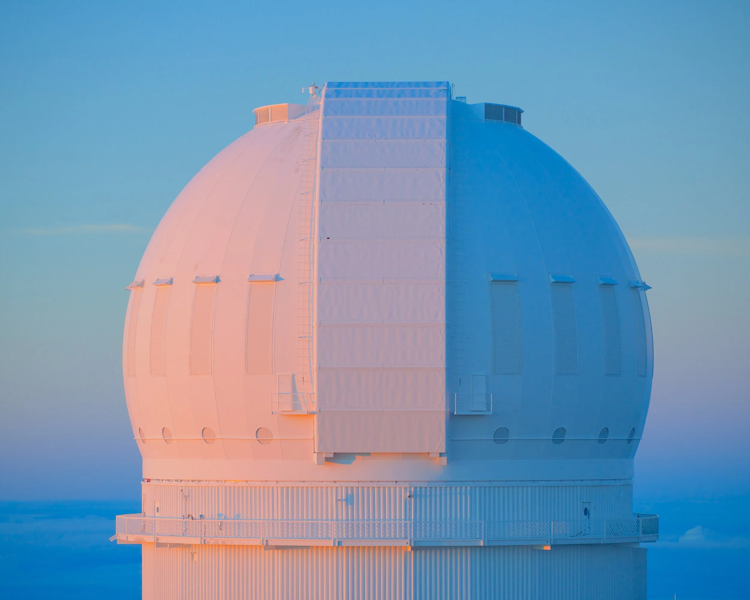 A large white observatory dome with a metal railing around the base, set against a clear sky at sunset or sunrise.