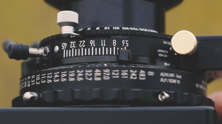 Close-up of a camera lens focus ring with measurement markings and a gold-colored knob, part of a professional camera or film camera.