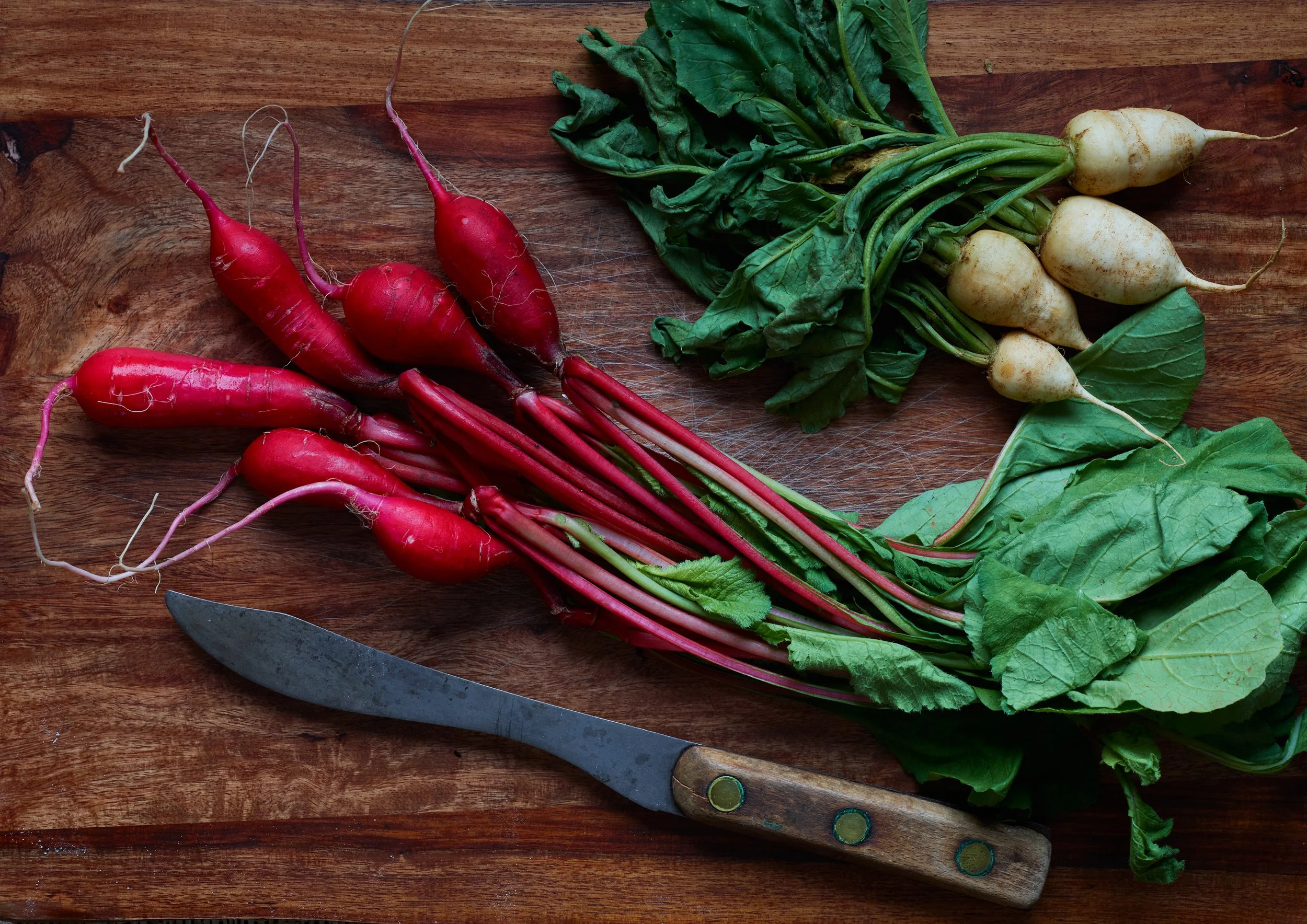 Bunch of fresh red radishes and white turnips on a wooden cutting board with a knife.