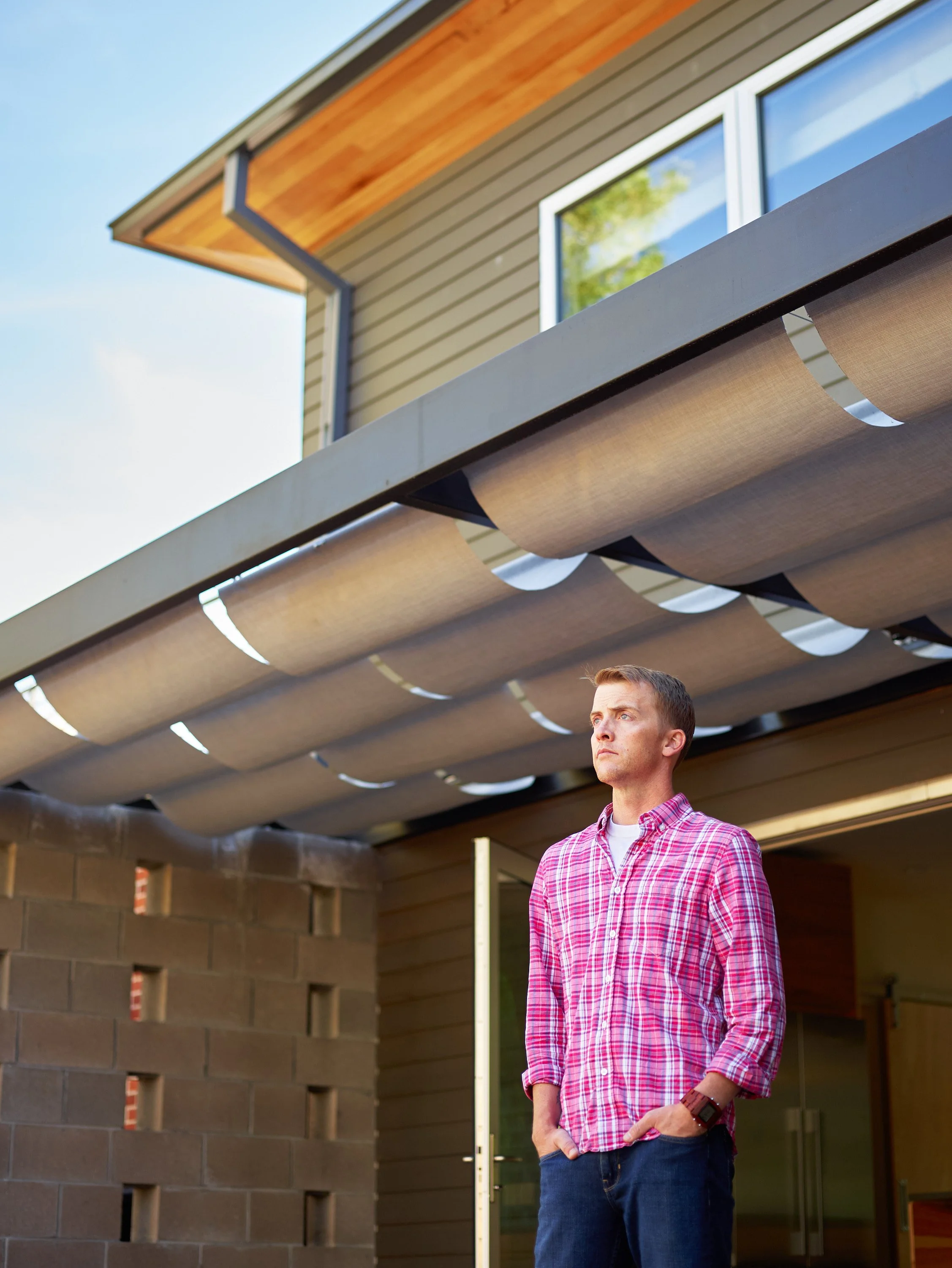 A man standing outside a modern house with a beige exterior, large windows, and a wooden roof overhang. He is wearing a red and white checkered shirt, blue jeans, and a smartwatch, looking thoughtfully into the distance.