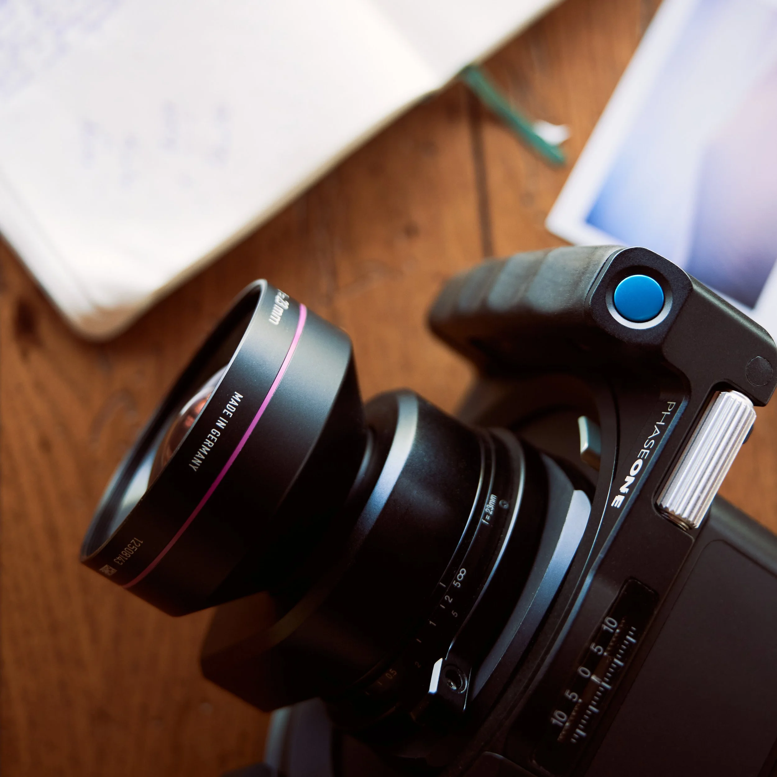 Close-up of a Phase One medium format camera with a large lens, placed on a wooden surface, with a laptop and a tablet partially visible in the background.