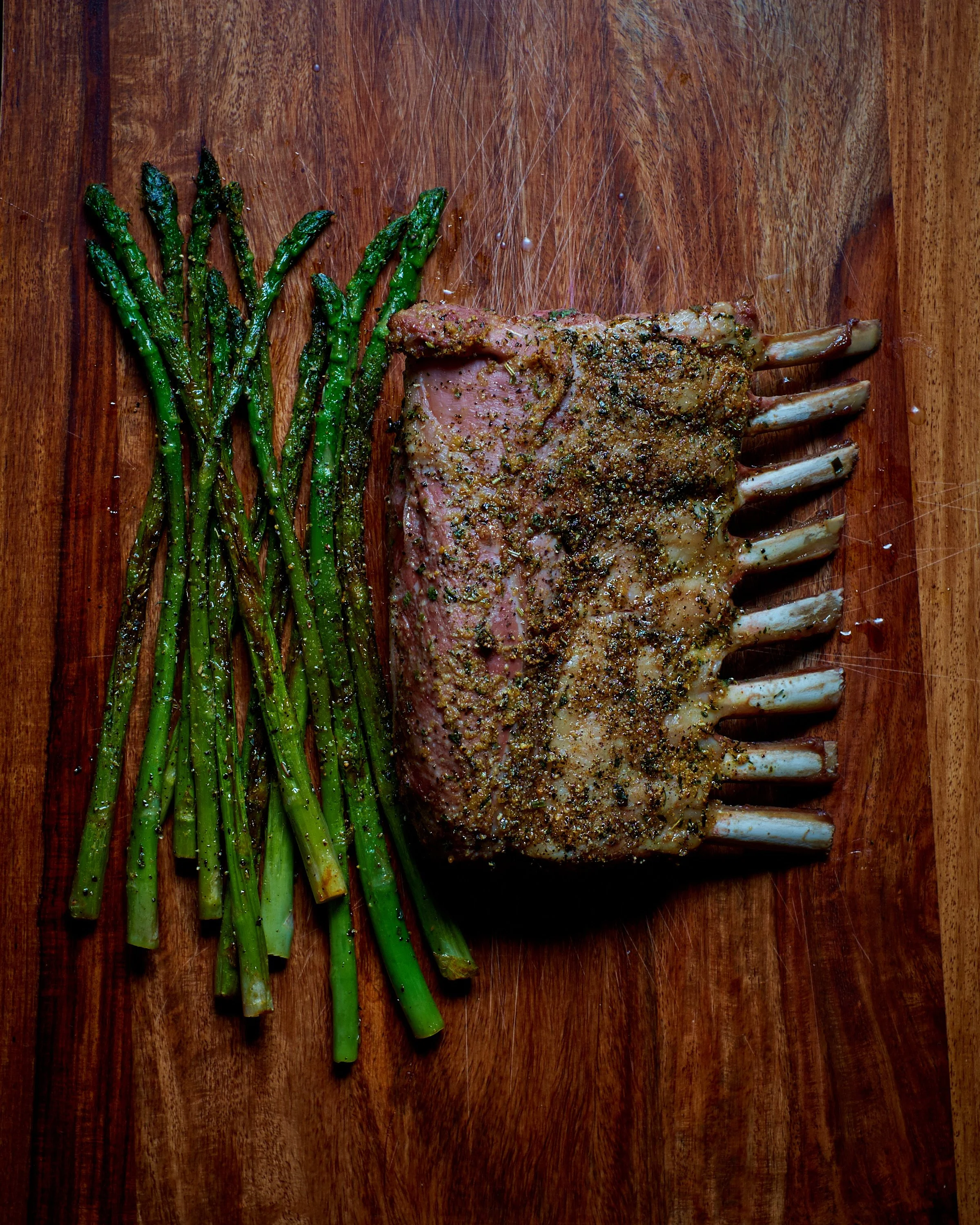 A wooden cutting board with seasoned rack of lamb and cooked green asparagus spears.