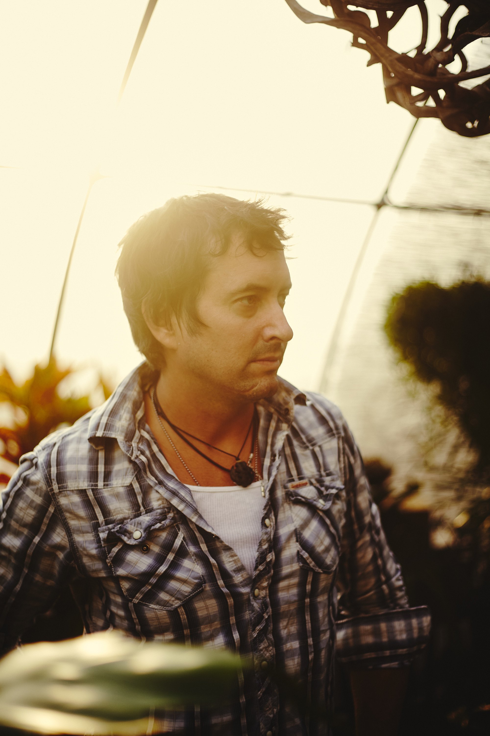 A man with dark, wavy hair wearing a plaid shirt and layered necklaces, standing outdoors at sunset with sunlight shining behind him.